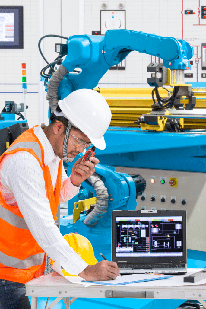 Engineer using a laptop computer for operation of factory automation, specifically an automatic robotic hand machine tool, in an industrial manufacturing facility.